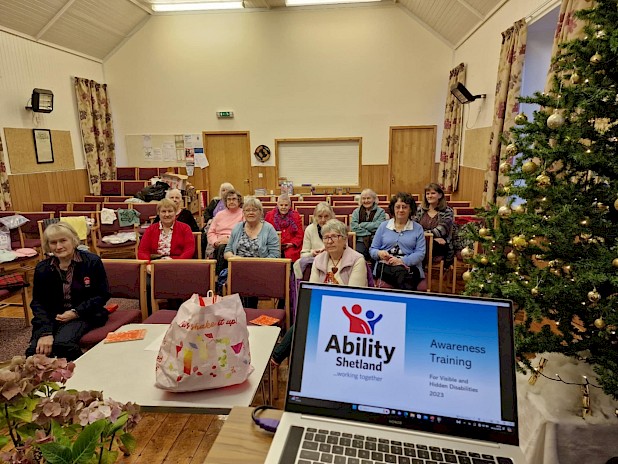 12 ladies from the Church of Scotland Guild sitting in a church hall looking towards the camera. A laptop is open at the front of the hall with a screen showing that says Awareness Training